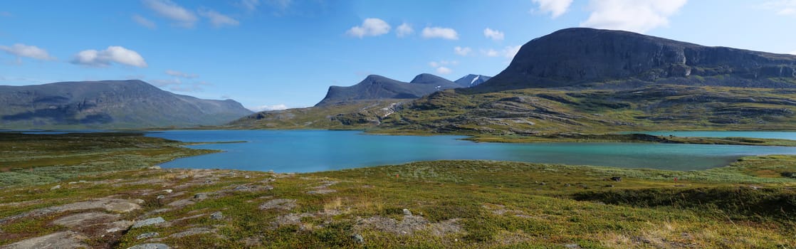 Lapland nature landscape wide panorama of blue glacial lake Allesjok near Alesjaure, birch tree forest, snow capped mountains. Northern Sweden, at Kungsleden hiking trail. Summer sunny day.