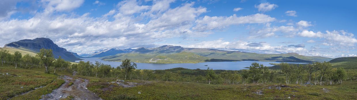 Panoramic landscape with beautiful river Lulealven, snow capped mountain, birch tree and footpath of Kungsleden hiking trail near Saltoluokta, north of Sweden, Lapland wild nature. Summer blue sky.