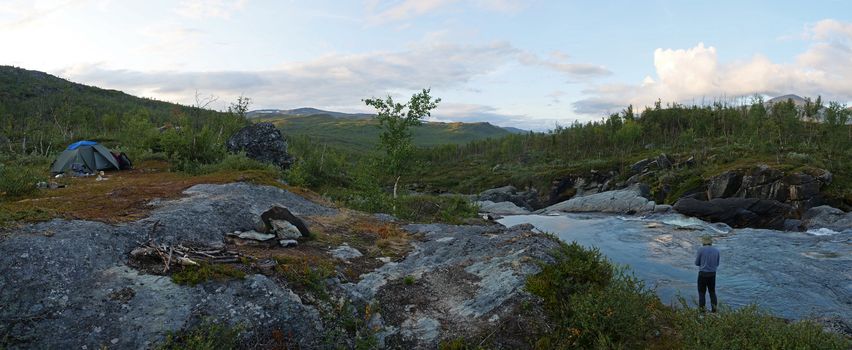 Wide panoramic Lapland landscape with with angler fisherman and small tent and rapids of river Kamajakka, rock, birch trees and mountains at Rovvidievva sami village, Northern Sweden, Summer evening.