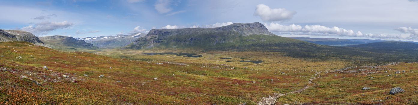Panoramic landscape of wild nature in Sarek national park in Sweden Lapland with snow capped mountain peaks, river and lake, birch and spruce tree forest. Early autumn colors, blue sky white clouds