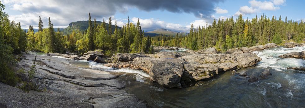Beautiful wide panoramic landscape with wild glacier river Kamajokk, boulders and spruce tree forest in Kvikkjokk in Swedish Lapland. Summer sunny day, golden hour, dramatic clouds.