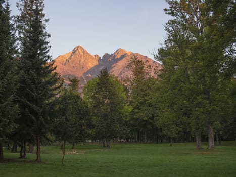 Tatra Mountains range colored pink and orange sunrise light , view from Tatranska Lomnice with green trees and grass. Early autumn panorama.