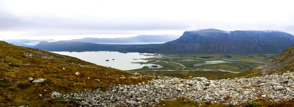 Panoramic view on meandering river delta at Rapadalen valley in Sarek national park, Sweden. Lapland mountains, rocks and birch trees. Early autumn colors, moody sky with clouds