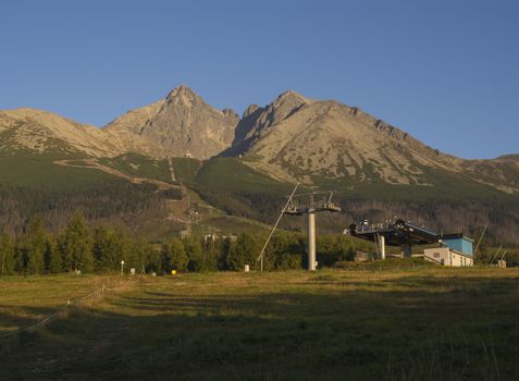 Tatra Mountains range gold colored sunrise light, ski lift at Tatranska Lomnice, start with green trees and grass. Early autumn panorama