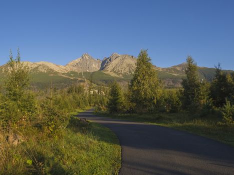 Tatra Mountains range with asphalt road curve, golden hour light, way from Tatranska Lomnice to Start with green trees and grass. Early autumn panorama.