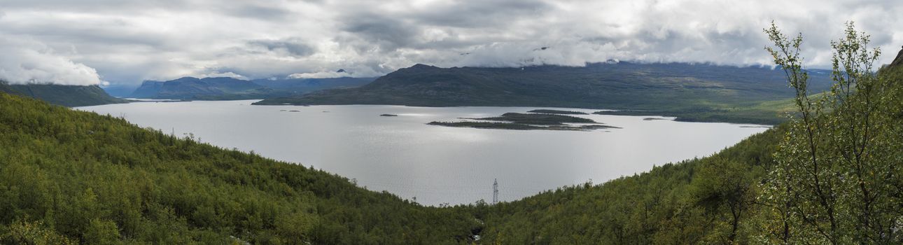 Panoramic landscape of Akkajaure lake at Kungsleden hiking trail, green mountains and birch tree forest in mist and clouds.