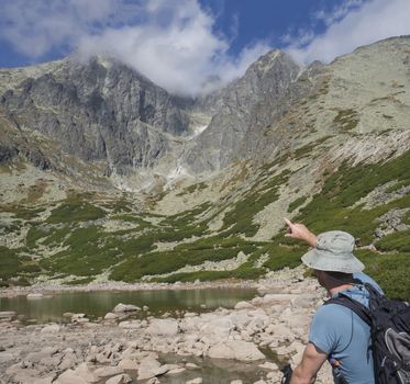 Young man hiker looking and point to mountain Peak Lomnicky stit 2 634 m covered in clouds at Summer, the highest mountain peaks in the High Tatras mountains of Slovakia Vysoke Tatry and mountain lake Skalnate pleso.