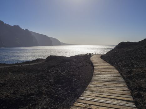 wooden planks path leading to the sea shore with view on hight lava cliffs los gigantes on Punta de Teno cape on west Tenerife canary island with lava rock blue sea and clear summer sky Contre-jour against daylight