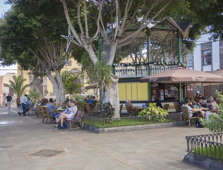 Spain, Canary islands,Garachico, December 19, 2017, group of people and tourist siting on the bench and fast food tables and relaxing in the public park with big trees
