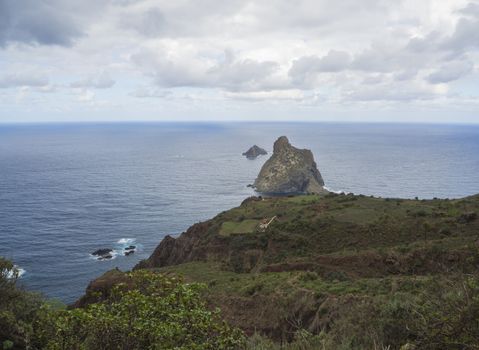 view on rock in the sea El Draguillo with green hills, farm house and dramatic blue sky white clouds in anaga mountain, tenerife  canary island spain