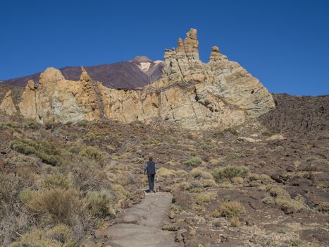 tourist people hikers on the path around famous pitoresque rock formation Roques de Garcia with view on colorful volcano pico del teide highest spanish mountain, clear blue sky background