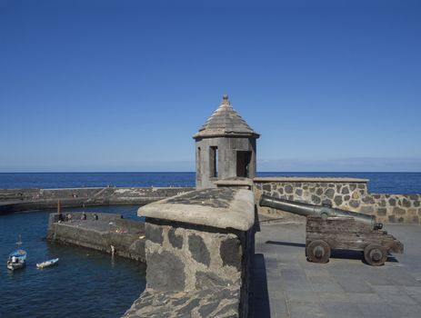 old copper cannon on paved sea front with embrasure tower, stone wall, sea horizon, fisherman boat and bathing people with clear blue sky background in Peurto de la Cruz, Tenerife