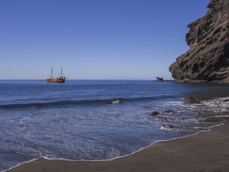 View on sea horizon with old Sailing ship from black sand beach Playa De Masca with big  cliffs and blue sky background