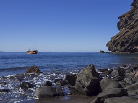 View on sea horizon with old Sailing ship from black sand beach Playa De Masca with big stones, cliffs and blue sky background