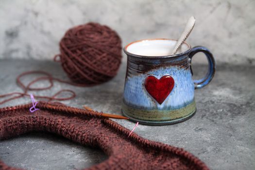 Brown sweater, knitting process, with ceramic cup with red heart on concrete background