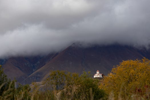 Kazbegi region of Georgia, snow capped mountains and rugged terrain. White Orthodox church with red roofs near Almasiani Georgia with mountains and dark clouds in the background, very atmospheric and darkHigh quality photo