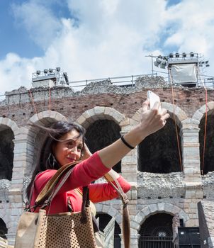 Young woman tourist, with her fashion cork bag, taking selfie photo in Verona (Italy). In background the famous Arena.