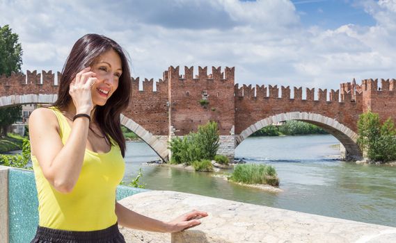Portrait of a young woman talking on phone. In the background the medieval stone bridge Scaligero built in 14th century near Castelvecchio, Verona (Italy). Shallow DOF.