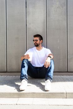 Bearded guy with sunglasses sitting on ground while holding a take away coffee