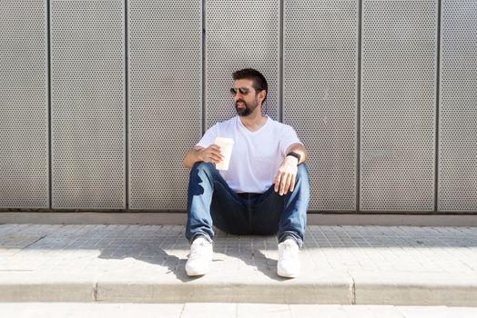 Bearded guy with sunglasses sitting on ground while holding a take away coffee