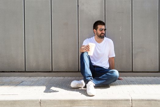 Bearded guy with sunglasses sitting on ground while holding a take away coffee