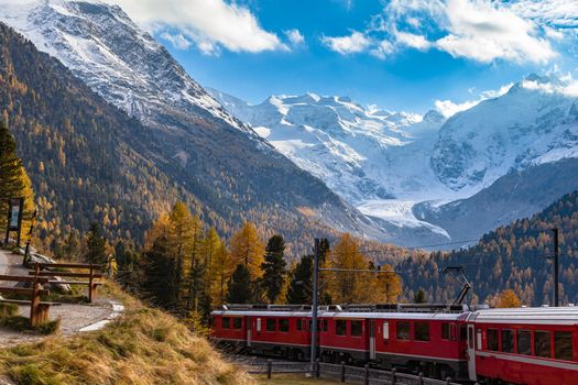 Stunning view of red Rhaetian train running under the Morteratsch Glacier in autumn with blue sky cloud, on sightseeing railway line Bernina Express, Canon of Grisons, Switzerland
