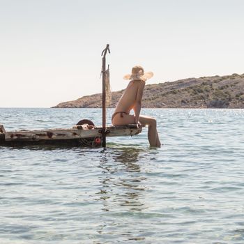 View of unrecognizable woman wearing big summer sun hat tanning topless and relaxing on old wooden pier in remote calm cove of Adriatic sea, Croatia.