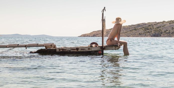 View of unrecognizable woman wearing big summer sun hat tanning topless and relaxing on old wooden pier in remote calm cove of Adriatic sea, Croatia.