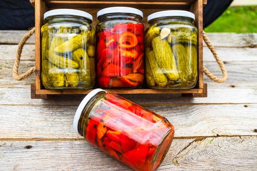 Glass jars with pickled red bell peppers and pickled cucumbers (pickles) isolated in wooden crate. Jars with variety of pickled vegetables. Preserved food concept in a rustic composition.