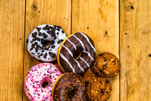 Colorful donuts and biscuits on wooden table. Sweet icing sugar food with glazed sprinkles, doughnut with chocolate frosting. Top view with copy space