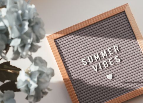 Letter board with words SUMMER VIBES and heart. Top view on flat lay board with quote with symbol of love and romance. Summer sun shining on white background and blue flowers in bloom.