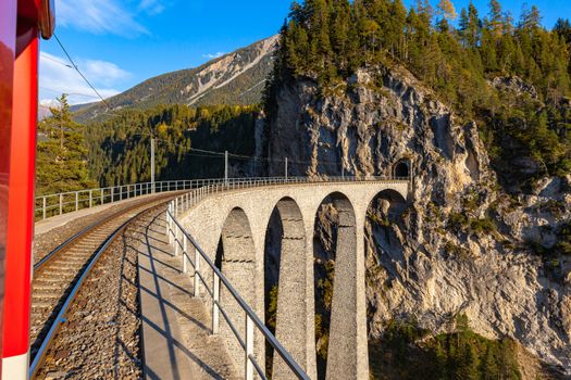 Travel with the red Rhaetian railway sightseeing train Bernina Express running over Landwasser Viaduct on sunny autumn day with blue sky cloud, Canon of Grisons, Switzerland
