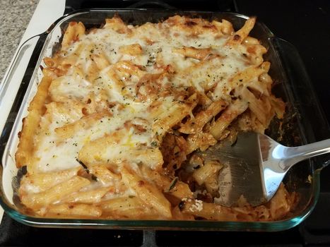 baked hot cheesy penne pasta in glass container on stove with spatula