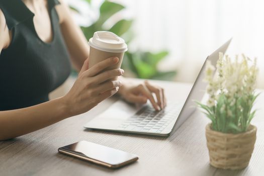 Young woman drinking coffee from disposable cup and using computer laptop on work desk.