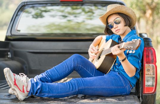 Young asian women short hair wear hat and sunglasses playing guitar ,sit on pickup truck in countryside Thailand