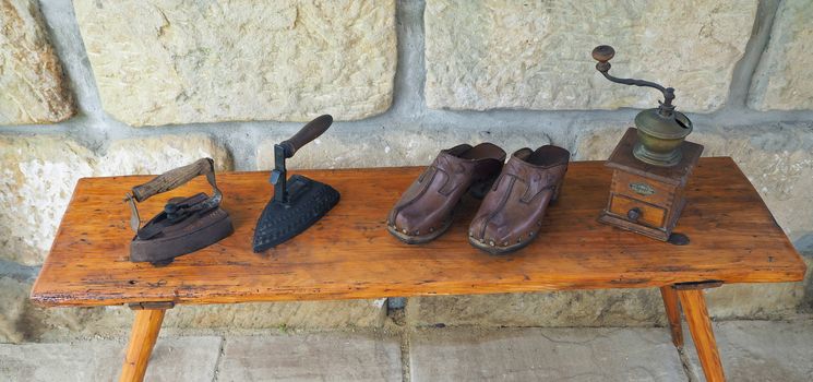 wo old rusty antique iron with wooden handle, hand coffee grinder and leather clogs on old wooden bench with sandstone wall background
