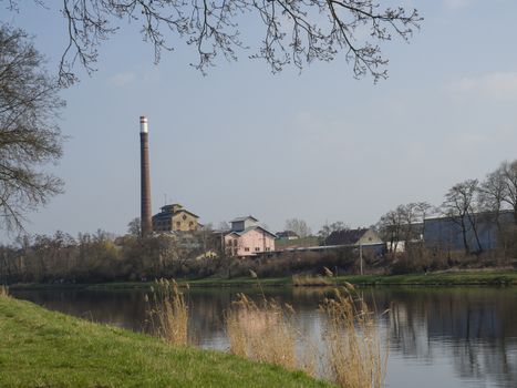 old historical factory building with chimney across the river elbe, lush green grass, tree and blue sky