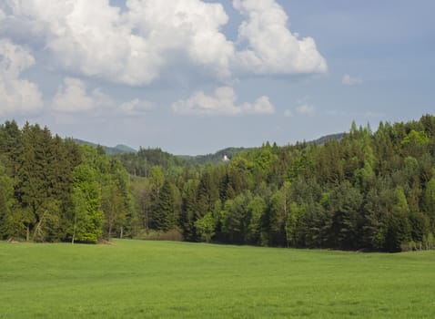 idyllic spring landscape with lush green grass meadow, fresh deciduous and spruce tree forest, small chapel and hills, blue sky white clouds background, horizontal, copy space