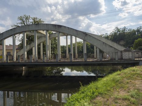 old arc bridge across Jizera river made concrete and metal with view on trees and village summer landscape, blue sky