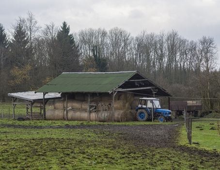 blue tractor with straw bale under porch and farming tools trees and green grass