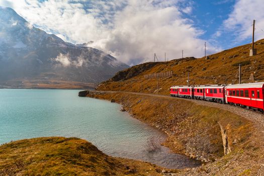 Beautiful view of  Rhaetian railway red train running on the lake side of Lago Bianco in autumn with blue sky cloud, on sightseeing railway line Bernina Express, Canon of Grisons, Switzerland