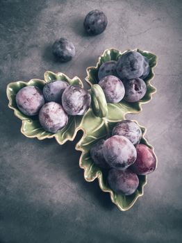 On the table in a ceramic dish are large ripe plums. Presented in close-up on a dark background.