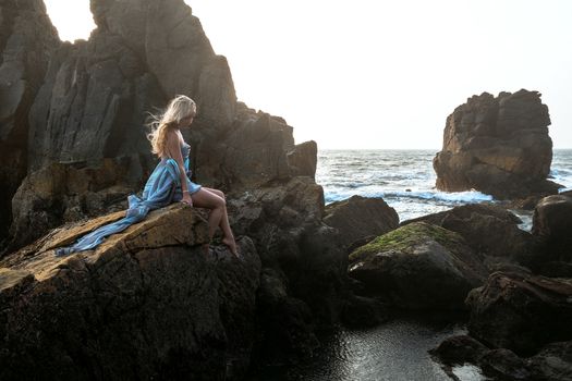 Beautiful European girl in a long dress sitting on the edge of a cliff and looking at the sea