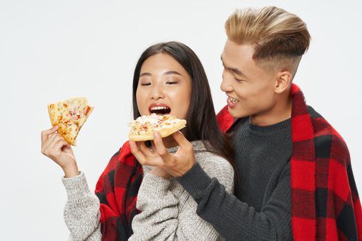A cheerful young Asian-looking couple covered in a plaid blanket is eating pizza