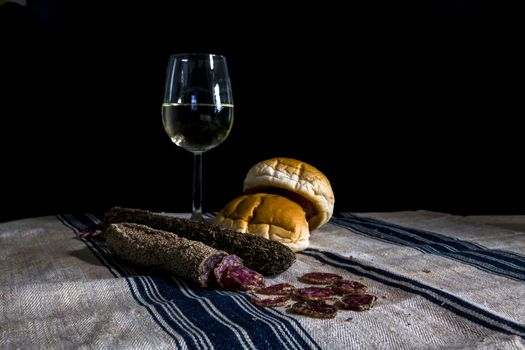still life table with a glass of white wine assorted dry sausages and bread on a flour cloth with blue stripes