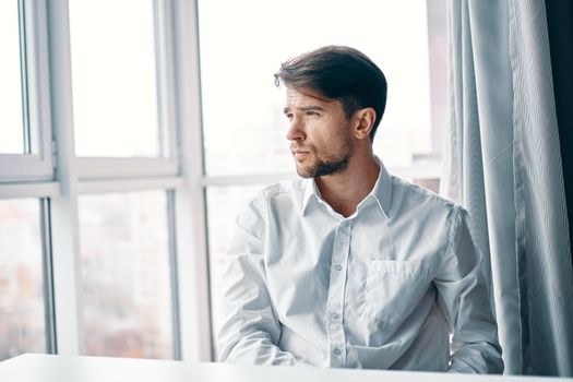 A puzzled man in a shirt looks out the window and sits near the table