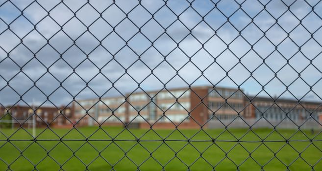A Typical High School Building And Sports Field Under A Cloudy Sky With Focus On The Security Fence In The Foreground