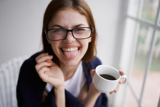 Cheerful woman with a cup of coffee shows tongue to the camera and glasses on her face