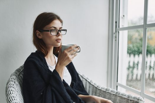 Pretty woman in glasses sits near the window with a cup of coffee in her hands