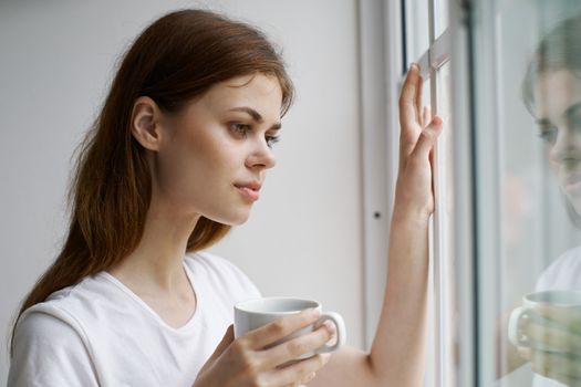 Beautiful woman near the window looks at nature and a cup of coffee in her hand